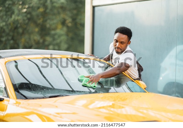 Car wash and cleaning at outdoors self service\
station. Shot of handsome bearded young guy cleaning sport car\
windscreen with a green microfiber cloth outdoors in summer sunny\
morning
