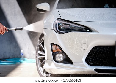 Car wash concept. Front view of white sports car covered with water droplet & beading on hood & bumper. Professional car detailing & cleaning service. Male working with high pressure water background.