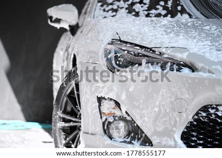 Car wash concept. Front view of white sports car covered with water, washing foam, and soap on hood & bumper. Professional car detailing & commercial cleaning service concept. Wet car background.