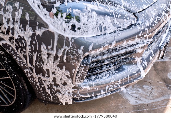 Car wash concept. Front view of white sports\
car covered with water, washing foam, and soap on hood &\
bumper. Professional car detailing & commercial cleaning\
service concept. Wet car\
background.