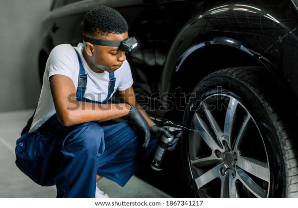 Car wash and\
detailing concept. Handsome African American male car wash worker\
in uniform, holding in hand special chemical cleaner solution and\
spraying it on car wheel\
rim.