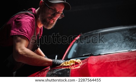 Car Wash Expert Using a Microfiber Cloth to Dry Up a Red Modern Sportscar. Adult Man Cleaning Away Dirt, Preparing an American Muscle Car for Detailing. Creative Cinematic Photo with Sport Vehicle