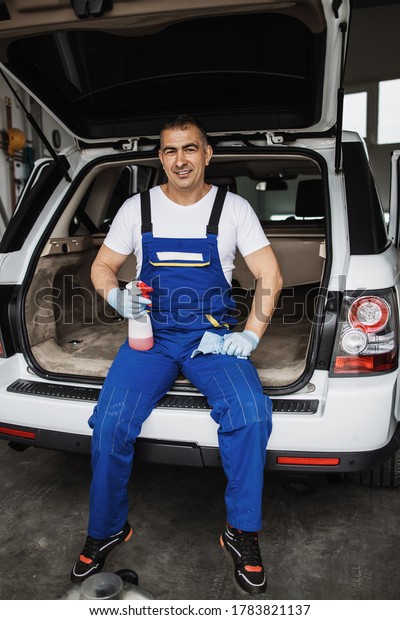 Car wash service worker sitting and posing in\
front of luxurious vehicle. Car cleaning, polishing and valeting\
service.