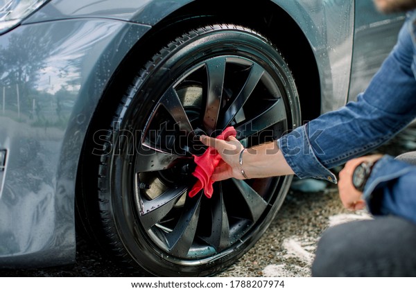 Car washing and\
detailing photo, car wash outdoors. Cropped image of young man,\
washing modern car alloy wheel on a car wash, using red microfiber\
cloth and special cleaner