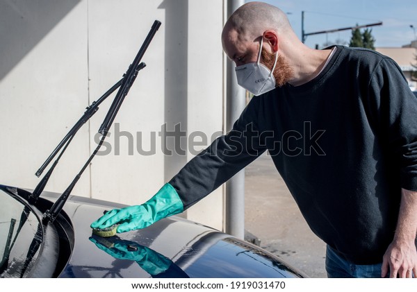 Car\
washing. Young man cleaning his car using a sponge. He is wearing a\
KN95 ffp2 mask against SARS-CoV-2 and a green\
glove