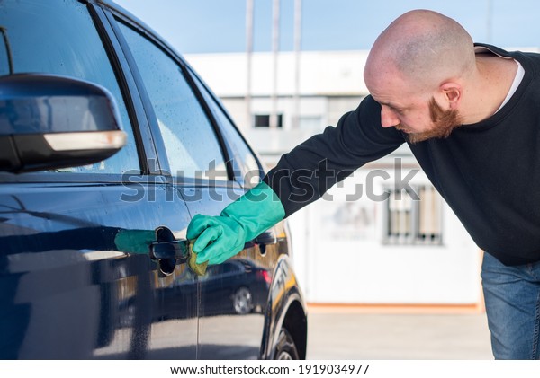 Car washing. Young man cleaning his car using\
a sponge. He is wearing a green\
glove