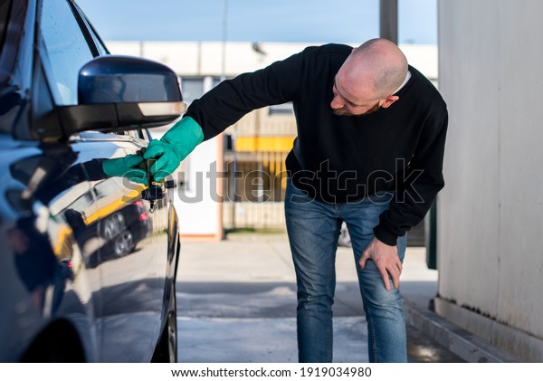 Car washing. Young man cleaning his car using\
a sponge. He is wearing a green\
glove