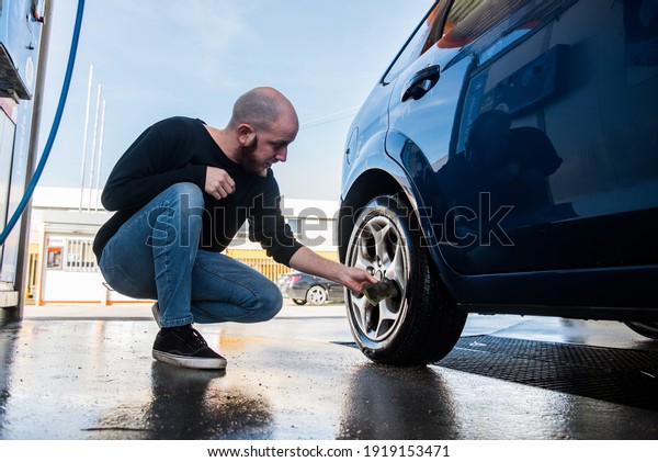 Car washing. Young man cleaning the dirt
of a tire of his car using a little
sponge.