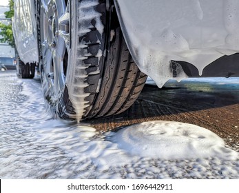 Car Wheel And Bumper Covered With Thick Fluffy Foam Dripping On Asphalt At A Do It Yourself Car Wash. Avoid Personal Contact And Wash Alone.