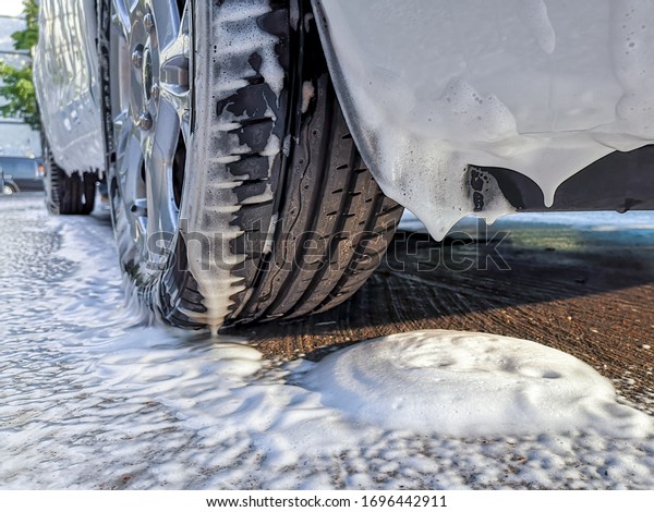 Car wheel and bumper covered with thick fluffy\
foam dripping on asphalt at a do it yourself car wash. Avoid\
personal contact and wash\
alone.