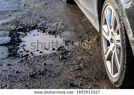 Car wheel close-up near a pothole on the road. Broken asphalt on the  roads. Spring and autumn problems for drivers. Potholes, water pits on a bad road.