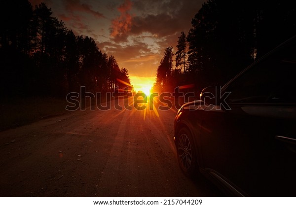 A car wheel travels along a dirt road on a journey\
to the horizon, against the backdrop of a bright sunset sky with\
rays of sun and black silhouette of trees. Freedom of movement.\
Side view rear