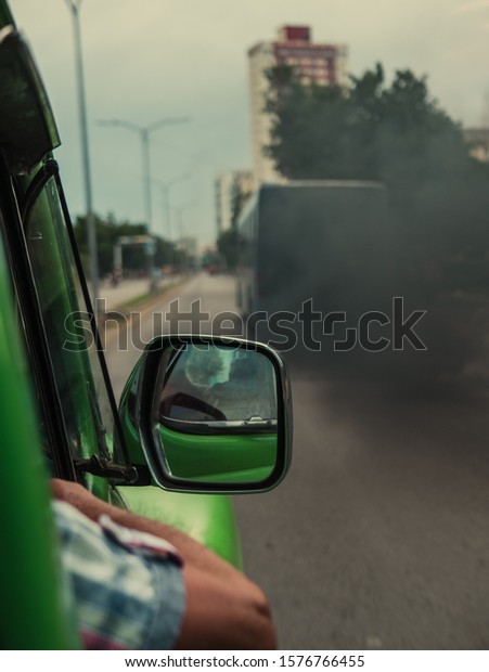 Car window with\
air pollution, expeled by an urban bus in city street, man with arm\
out of a green old car, side mirror. global warming with CO2\
emissions by combustion\
engine.