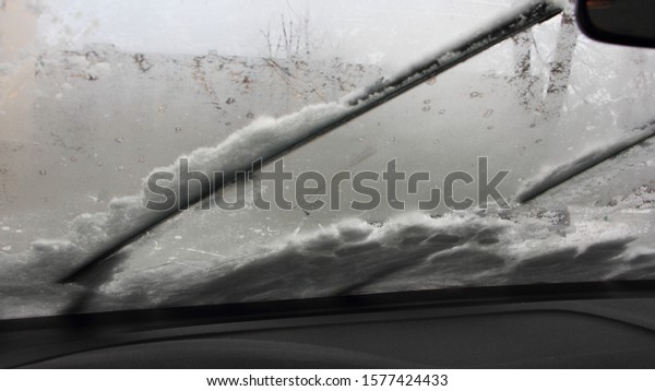 Car wiper blades clean the iced windshield from\
snow, inside view - winter cold start, snowstorm, snowfall, safety\
winter driving