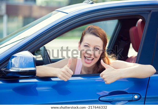 Car. Woman driver happy smiling showing thumbs\
up coming out of blue car side window on outside parking lot\
background. Beautiful young woman happy with her new vehicle.\
Positive face expression