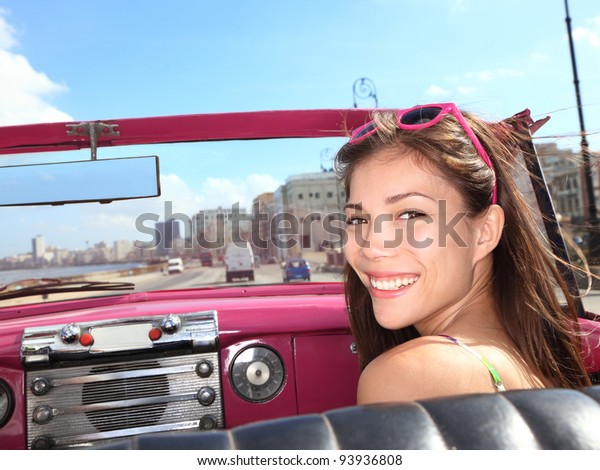 Car\
woman smiling happy on passenger seat in pink vintage convertible\
car. Young mixed race Asian / Caucasian female model during cuba\
vacation. Driving on Malecon waterfront, Havana,\
Cuba