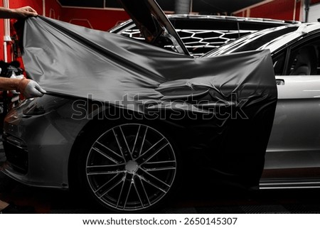 Car wrapping process using black matte vinyl stretched into place. Careful application and tensioning of black vinyl avoids surface damage or bubbling. Matte black vinyl is applied across car hood