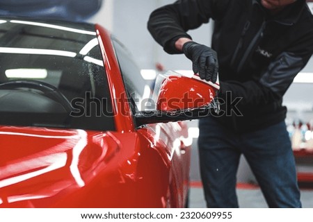 Car wrapping specialist in protective gloves wrapping red car's side mirror with transparent foil to protect the paint. High quality photo