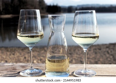 Carafe with white wine and two glasses of white wine on the left and right side. Beautiful landscape with a lake in the background, Allgäu, Bavaria
