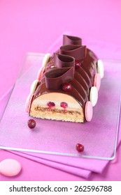 Caramel, Cherry and Praline Yule Log Cake glazed with chocolate mirror glaze, decorated with chocolate loops, cherries and french macarons, on a transparent cutting board and bright purple background.