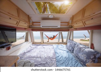 Caravan motorhome parked on the beach in front of the blue sea. Man laying in hammock and working with tablet on beach, viewing from inside caravan.
