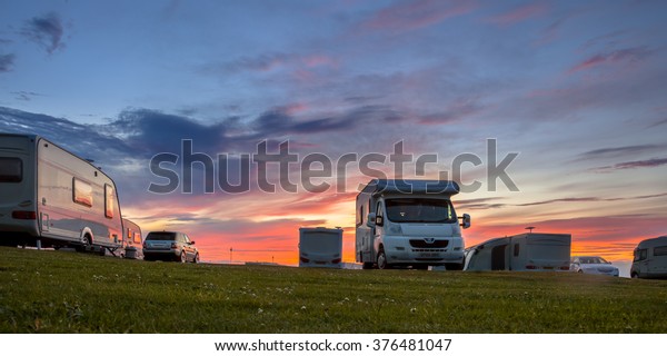 Caravans and cars parked on a grassy campground in\
summer under beautiful\
sunset
