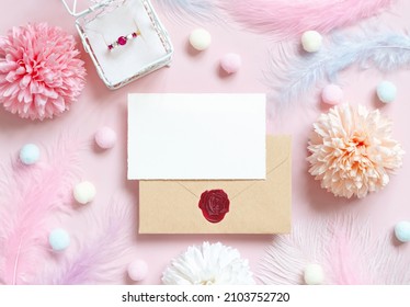 Card and envelope near pastel flowers, pom-poms, feathers and ring in a gift box on pink top view. Romantic scene with blank envelope and place for text flat lay. Valentines, Spring or girlish concept