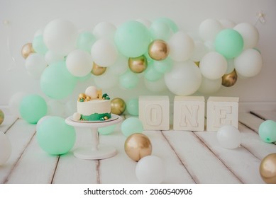 Card. Stylish photo zone in light colors with a cake and balloons in white mint and gold, wooden cubes with the inscription one. Light background white wood floor. Сake smash concept.