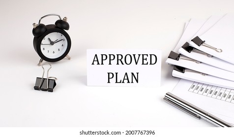 Card with text APPROVED PLAN on a white background, near office supplies and alarm clock. Business