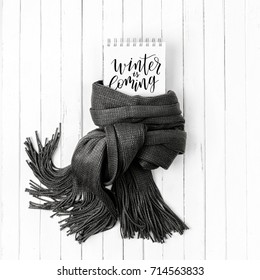 Card with word Winter is Coming wrapped in scarf on a white wooden background. Flat lay, top view