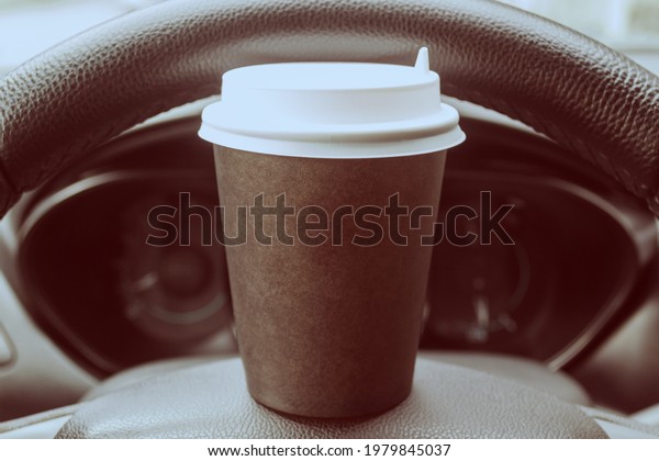 A cardboard\
cup of coffee on the steering wheel of a car. Rest stop on the\
road. Have a quick snack in the\
car.