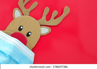 Cardboard Cutout Of A Red-nosed Reindeer Peeking While Wearing A Face Mask. Covid During Christmas Season Concept. Red Background With Copy Space.