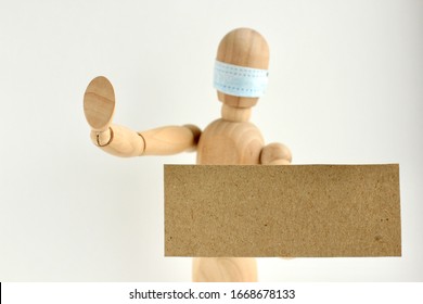 Cardboard Sign With Space For Text In The Hands Of A Wooden Doll Or Mannequin In A Medical Mask. Concept Of Anti Racism, Anti Xenophobia, The Social Situation Around The Coronavirus Or Covid-19