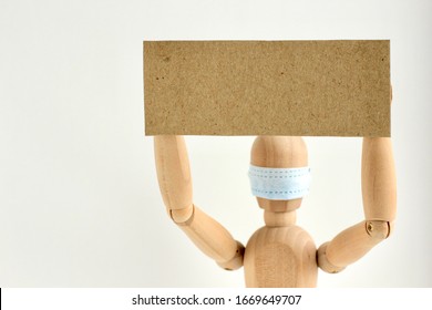 Cardboard Sign With Space For Text In The Hands Of A Wooden Doll Or Mannequin In A Medical Mask. Concept Of Anti Racism, Anti Xenophobia, The Social Situation Around The Coronavirus Or Covid-19