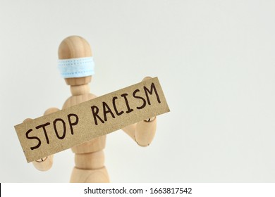 Cardboard Sign With Text Stop Racism In The Hands Of A Wooden Doll Or Mannequin In A Medical Mask. Concept Of Anti Racism, Anti Xenophobia, The Social Situation Around The Coronavirus Or Covid-19.