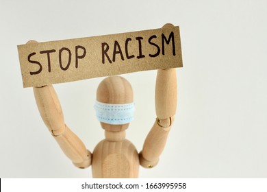 Cardboard Sign With Text Stop Racism In The Hands Of A Wooden Doll Or Mannequin In A Medical Mask. Concept Of Anti Racism, Anti Xenophobia, The Social Situation Around The Coronavirus Or Covid-19.