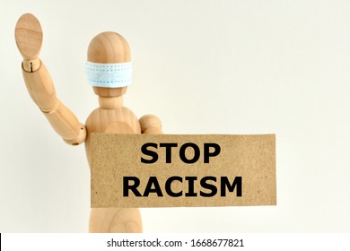 Cardboard Sign With Text Stop Racism In The Hands Of A Wooden Doll Or Mannequin In A Medical Mask. Concept Of Anti Racism, Anti Xenophobia, The Social Situation Around The Coronavirus Or Covid-19