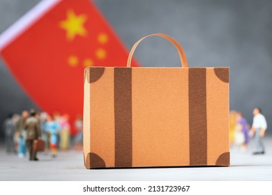 Cardboard Suitcase, Plastic Toy People And A Flag On An Abstract Background, A Concept On The Theme Of Moving Or Migration To China