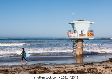 CARDIFF, CALIFORNIA/USA - APRIL 14, 2018:  A lone male surfer carries a surfboard past a lifeguard tower on San Elijo State Beach in Cardiff, California, located in San Diego County.