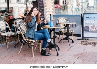 Cardiff, UK. March 2019. Young Lady Sitting At A Café Table, Talking On Her Cell Phone.