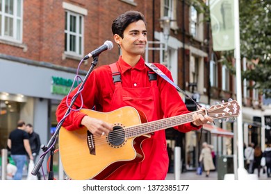 Cardiff, UK. October 2017. Young Asian Man Busking On His Guitar In The Hayes, Cardiff. He Is Wearing A Bright Red Shirt And Dungarees; And Has An Owl Earring.