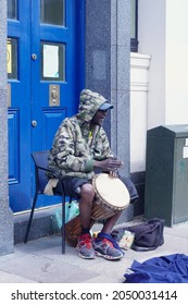 Cardiff, Wales, August 26 2021: A Happy Smiling Elderly Busker Is Sat In A Blue Doorway  In Cardiff City Centre Wearing A Hoodie Plays The Bongo Drums To Entertain Shoppers