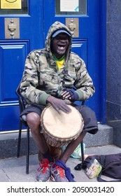 Cardiff, Wales, August 26 2021: A Happy Smiling Elderly Busker Is Sat In A Blue Doorway  In Cardiff City Centre Wearing A Hoodie Plays The Bongo Drums To Entertain Shoppers