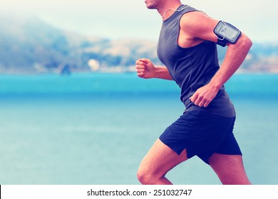 Cardio Runner Running Listening Smartphone Music. Unrecognizable Body Jogging On Ocean Beach Or Waterfront Working Out With Heart Rate Monitor App Device And Earphones In Summer.