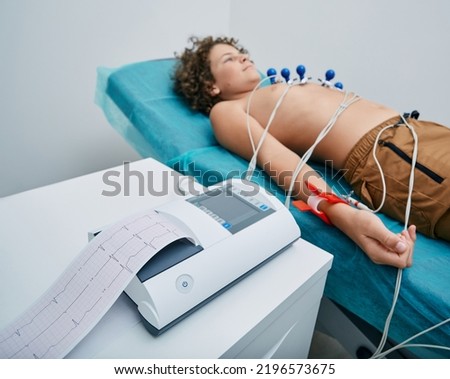 Cardiograph with ECG printout for teenager boy patient lying in medical bed with vacuum sensors. Heart electrocardiography for children