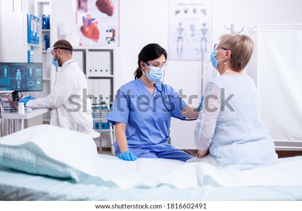 Cardiologist\
assistant listening heart of senior woman with stethoscope during\
consultation and wearing face mask against coronavirus outbreak.\
Medical examination for infections,\
disease.