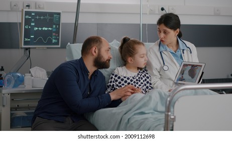 Cardiologist Woman Doctor Discussing Heart Cardiogram Explaining Recovery Treatment During Medicine Consultation In Hospital Ward. Little Child Lying In Bed After Sufffering Medical Cardiology Surgery