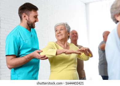 Care Worker Helping Elderly Woman To Do Sports Exercise In Hospital Gym.