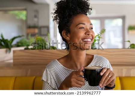 Carefree african american woman sitting in cafeteria drinking coffee while looking away. Black young woman drinking tea while thinking. Smiling girl relaxing and thinking while drinking cappuccino.