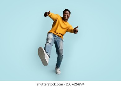 Carefree african american young man in casual outfit showing thumb ups and raising leg up, happy black guy posing on blue studio background, full length shot, panorama with copy space